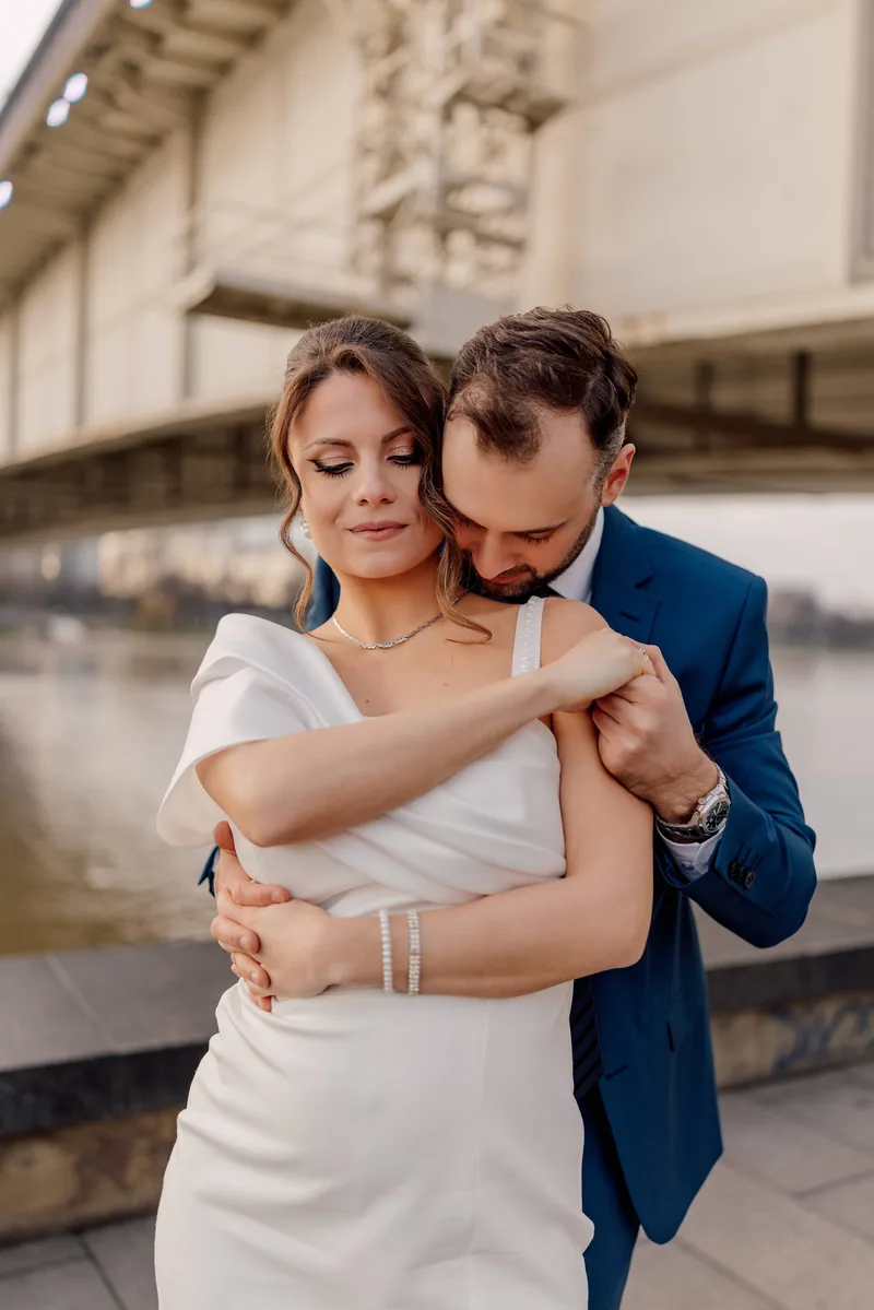 A groom embraces his bride from behind beside the river in warm golden light under a bridge.