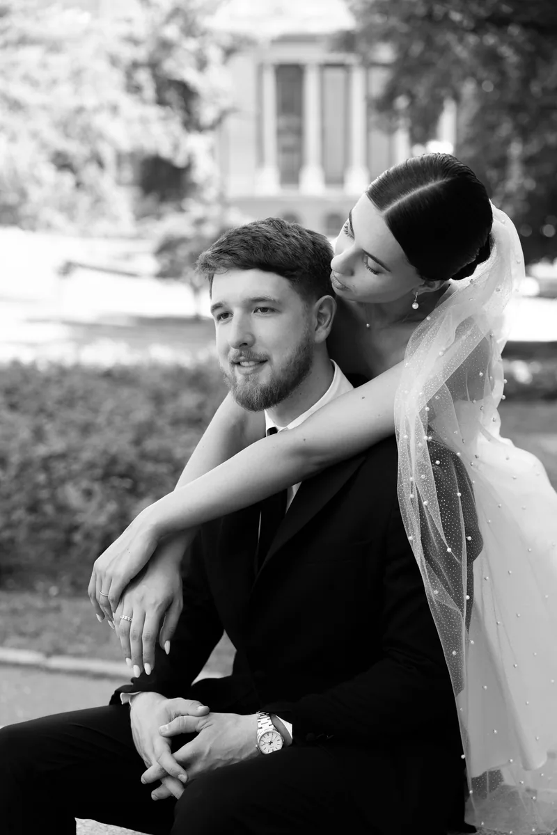 Black and white photo of a bride draping her arms around her seated groom and kissing near his cheek.