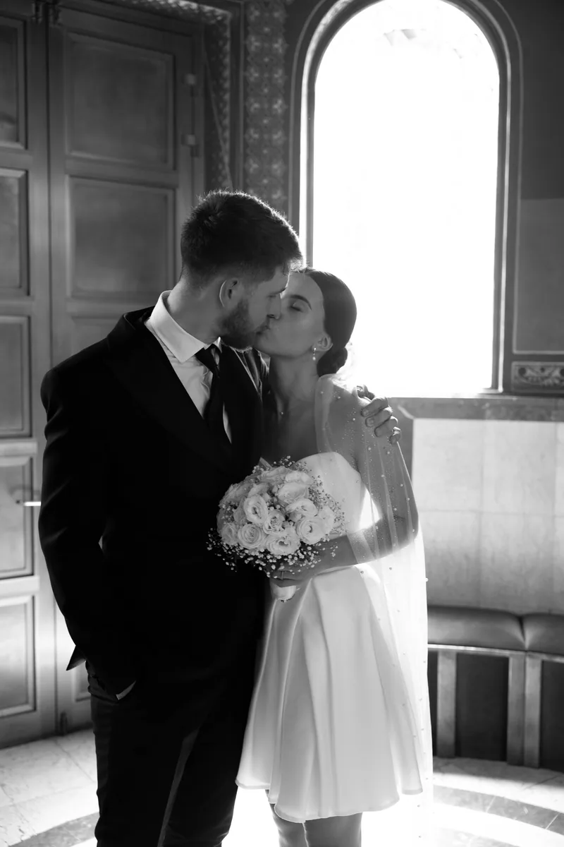 Black and white image of a couple kissing by a tall arched window, the bride holding a white rose bouquet.