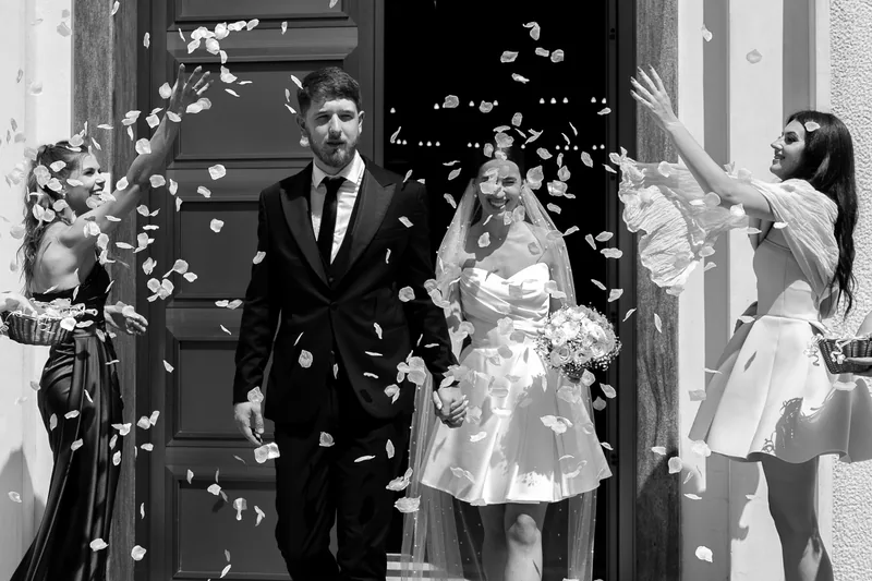 Black and white photo of newlyweds leaving a doorway as friends toss flower petals on both sides.