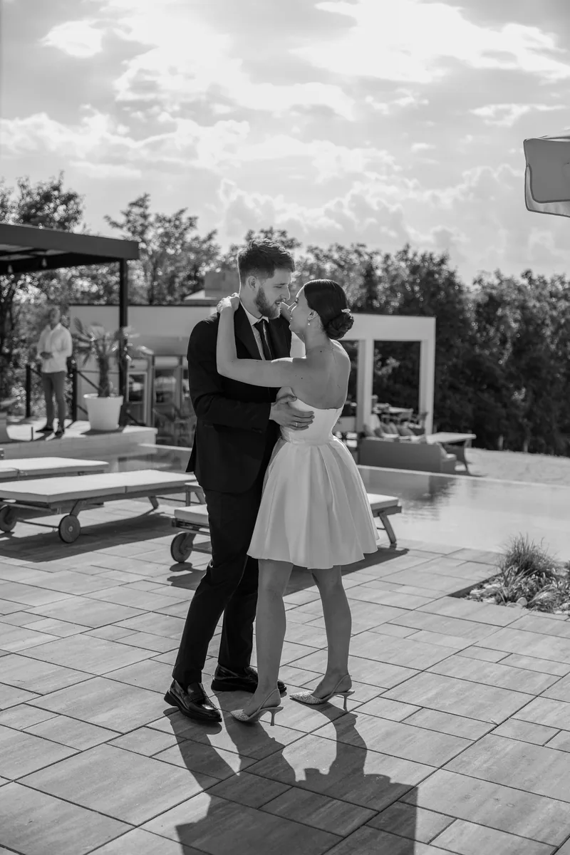 Black and white photo of a couple embracing in a dance on a sunlit terrace beside a swimming pool.