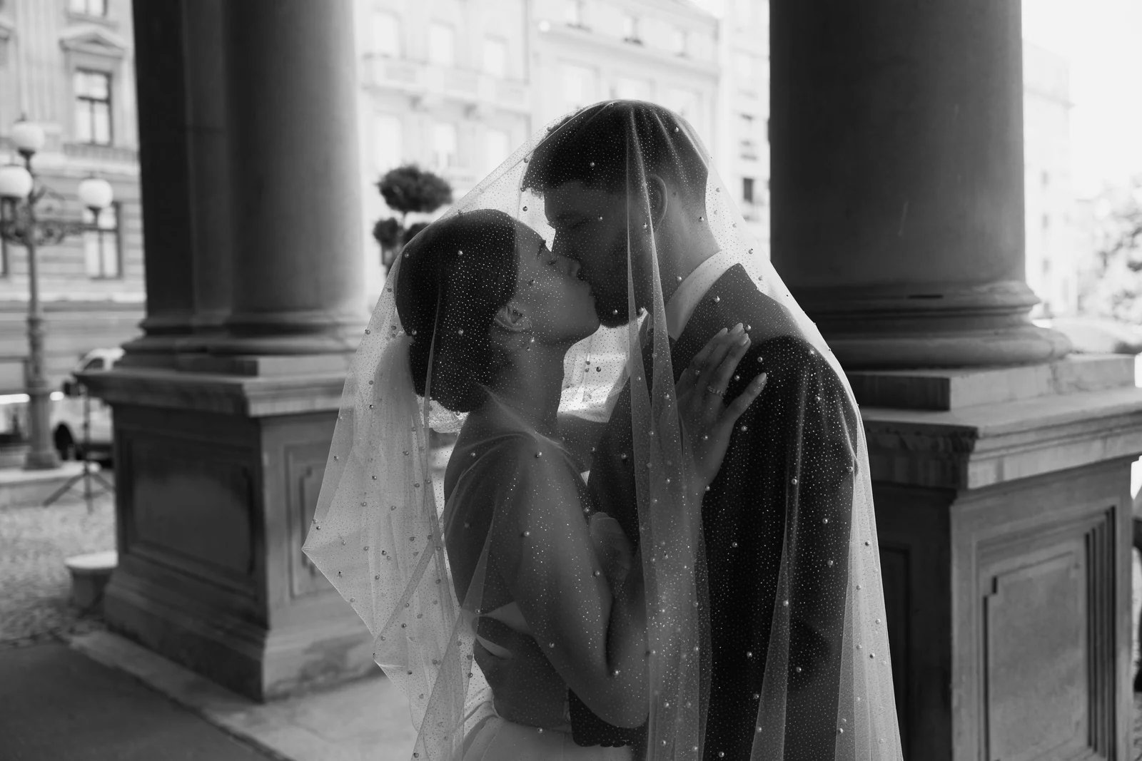 Black and white photo of newlyweds kissing beneath a sheer pearl veil between two tall stone columns.
