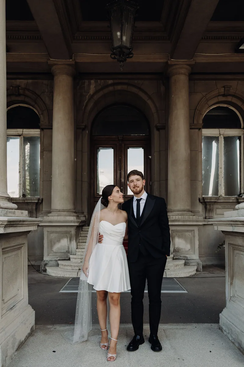 A bride gazes up at her groom as they stand together under an arched portico with tall stone columns.