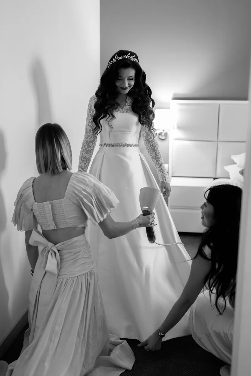 Bride in a lace gown and tiara is helped into her dress by two women before the wedding.