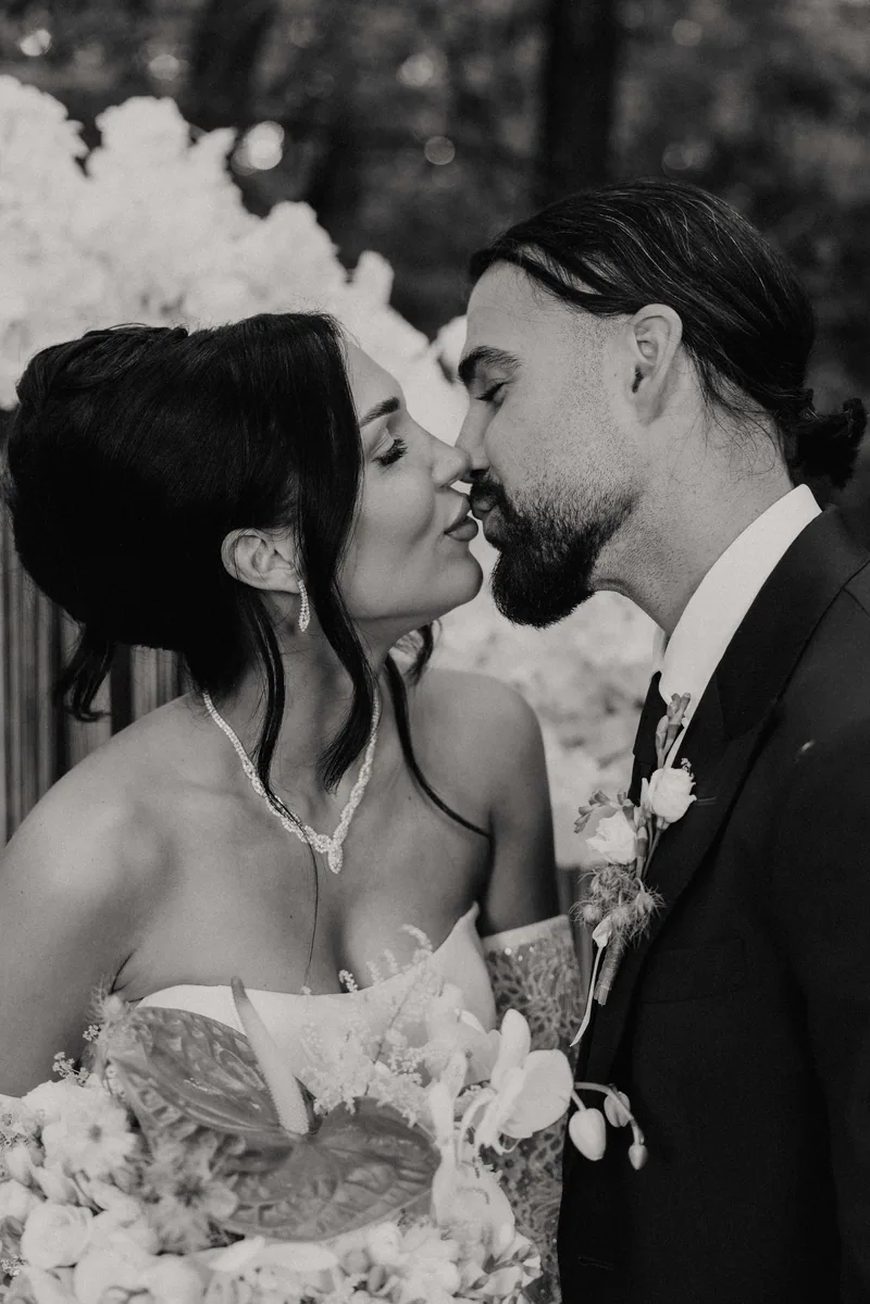 Black and white close portrait of a bride and groom leaning in to kiss beneath white orchids