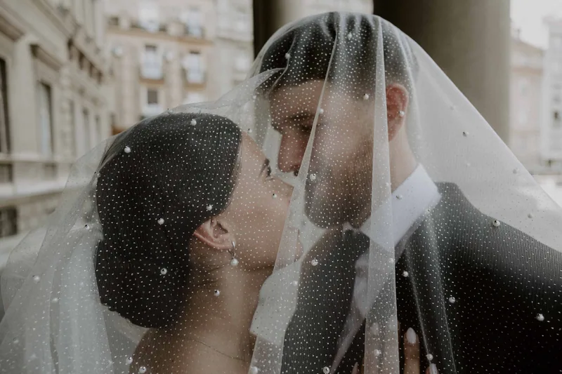 A bride and groom lean in close about to kiss beneath a pearl dotted veil that covers both their faces.