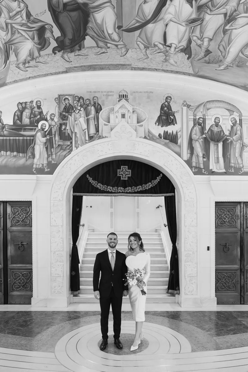 Bride and groom stand before an arched church portal painted with detailed frescoes.