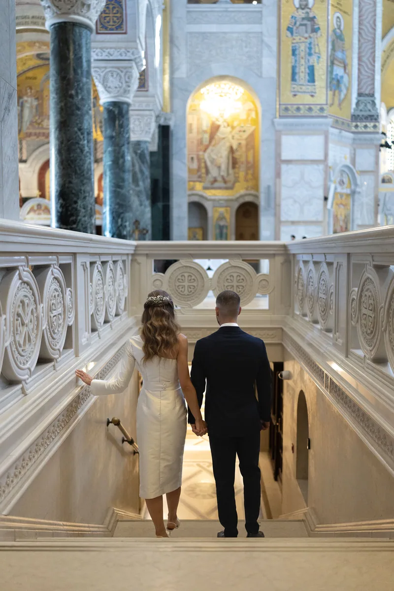 Bride and groom hold hands atop a white marble staircase framed by golden church mosaics.