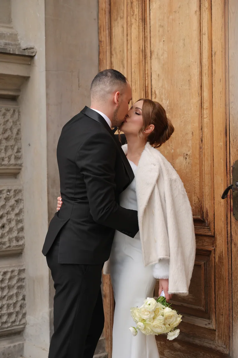 A bride in a white dress and cream fur coat kisses her groom in a black tuxedo before a tall aged wooden door.
