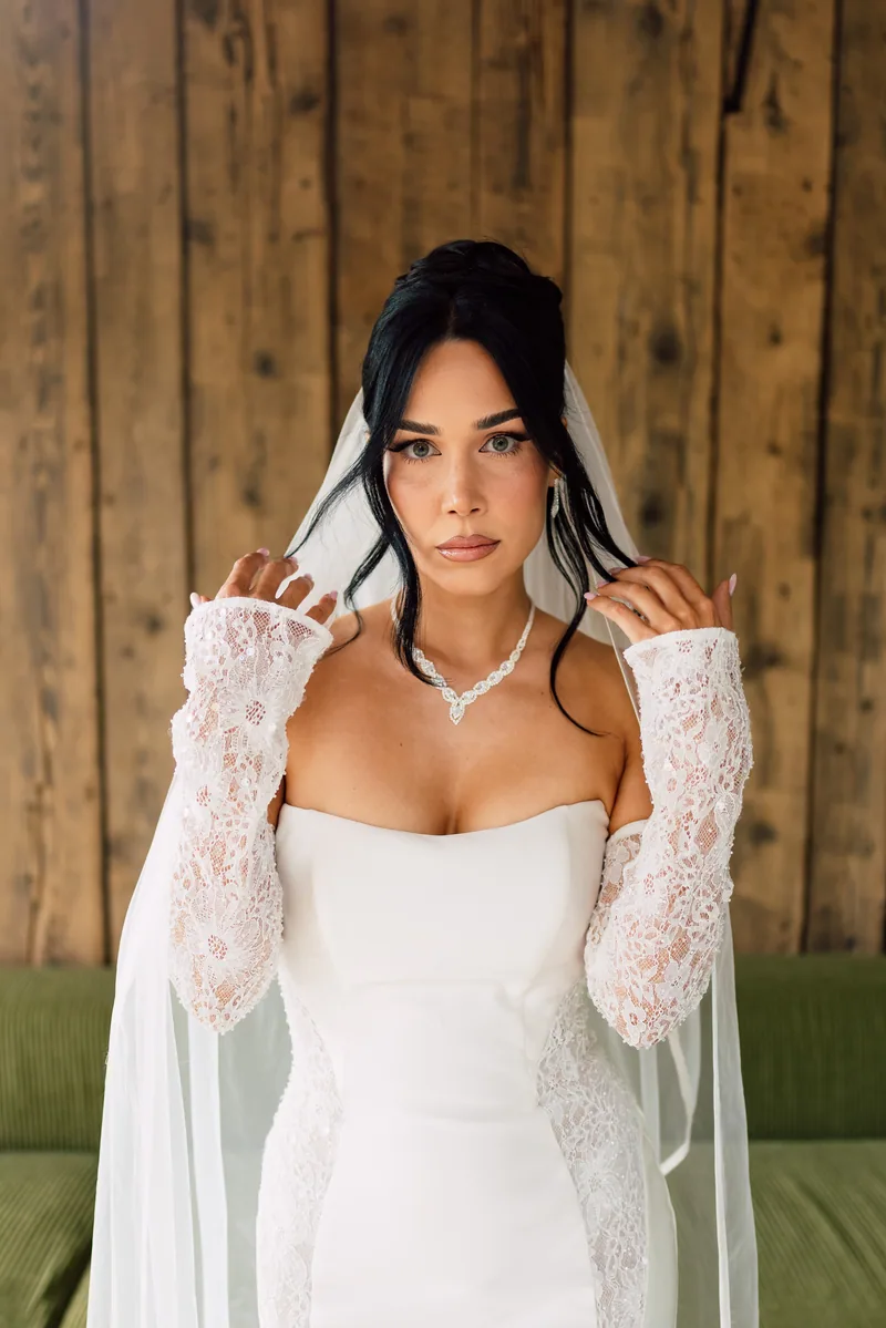 Bride lifting her sheer veil at both sides while wearing long lace gloves and a diamond necklace