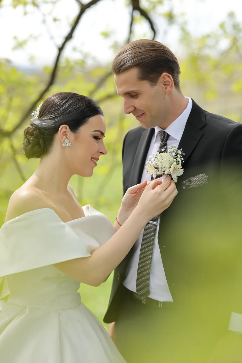 A bride adjusts the white boutonniere on her groom's lapel among soft green spring foliage.
