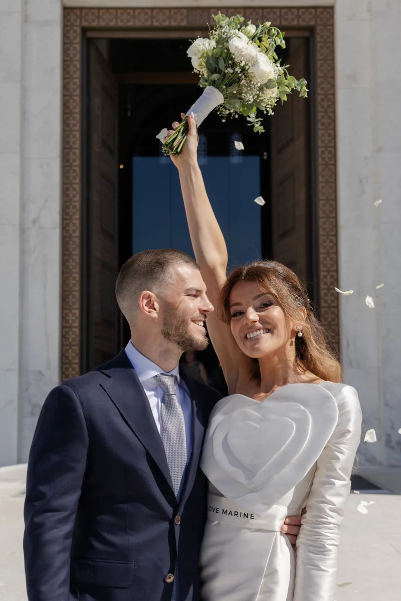 Bride lifts her bouquet in triumph as petals fall at the stone church doorway.