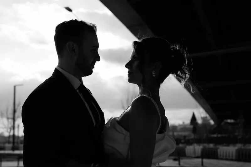 In black and white, a couple stand face to face in silhouette beneath a bridge against a cloudy sky.