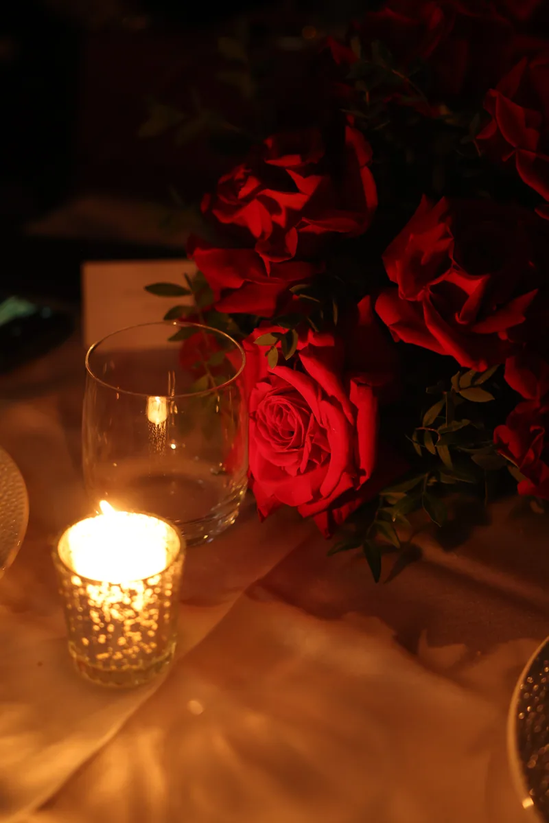 Warm candlelight glows over red roses and a votive candle on a reception table.