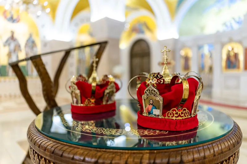 A pair of ceremonial wedding crowns sits on a glass table before a golden church nave.