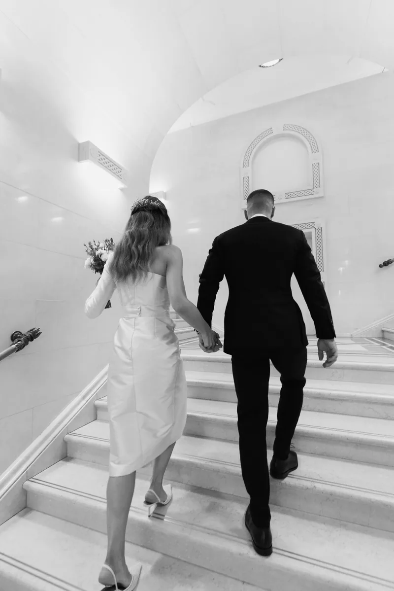 Couple seen from behind climb a curved white marble staircase, the bride carrying her bouquet.