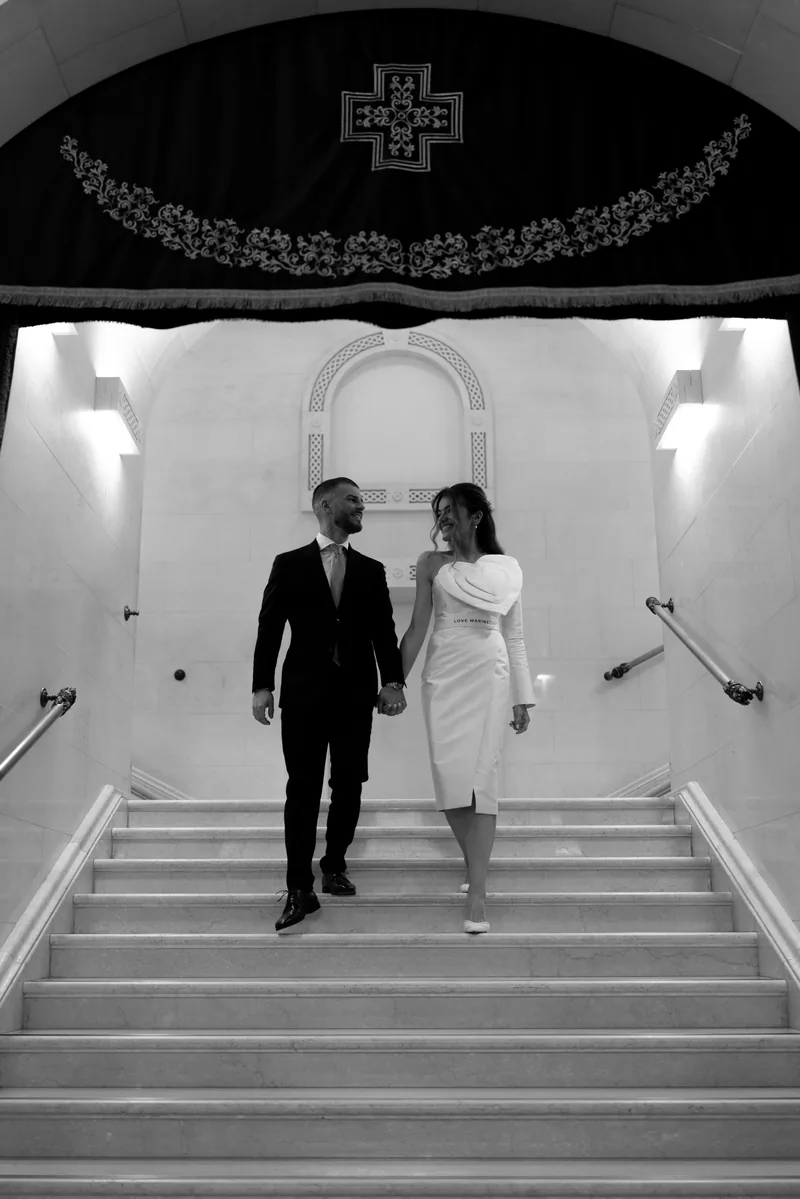 Black and white photo of a couple walking down grand stairs beneath an embroidered cross drape.