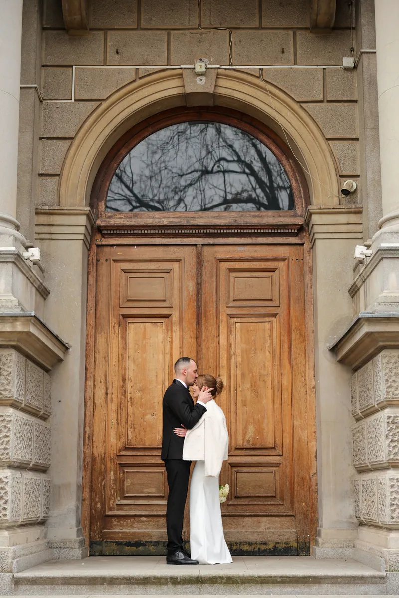 A newlywed couple embrace and kiss beneath a tall arched wooden doorway of a grand stone building.