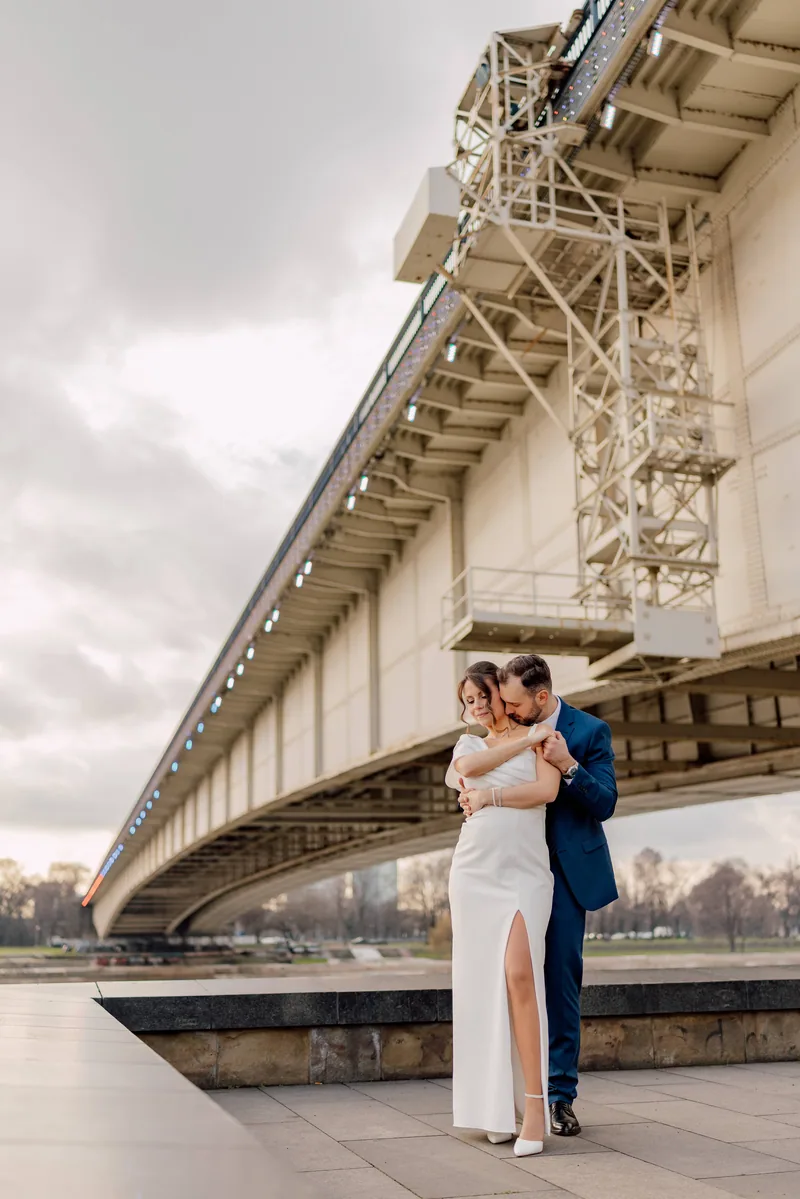 A couple embrace on a riverside walkway beneath a large illuminated bridge at dusk.