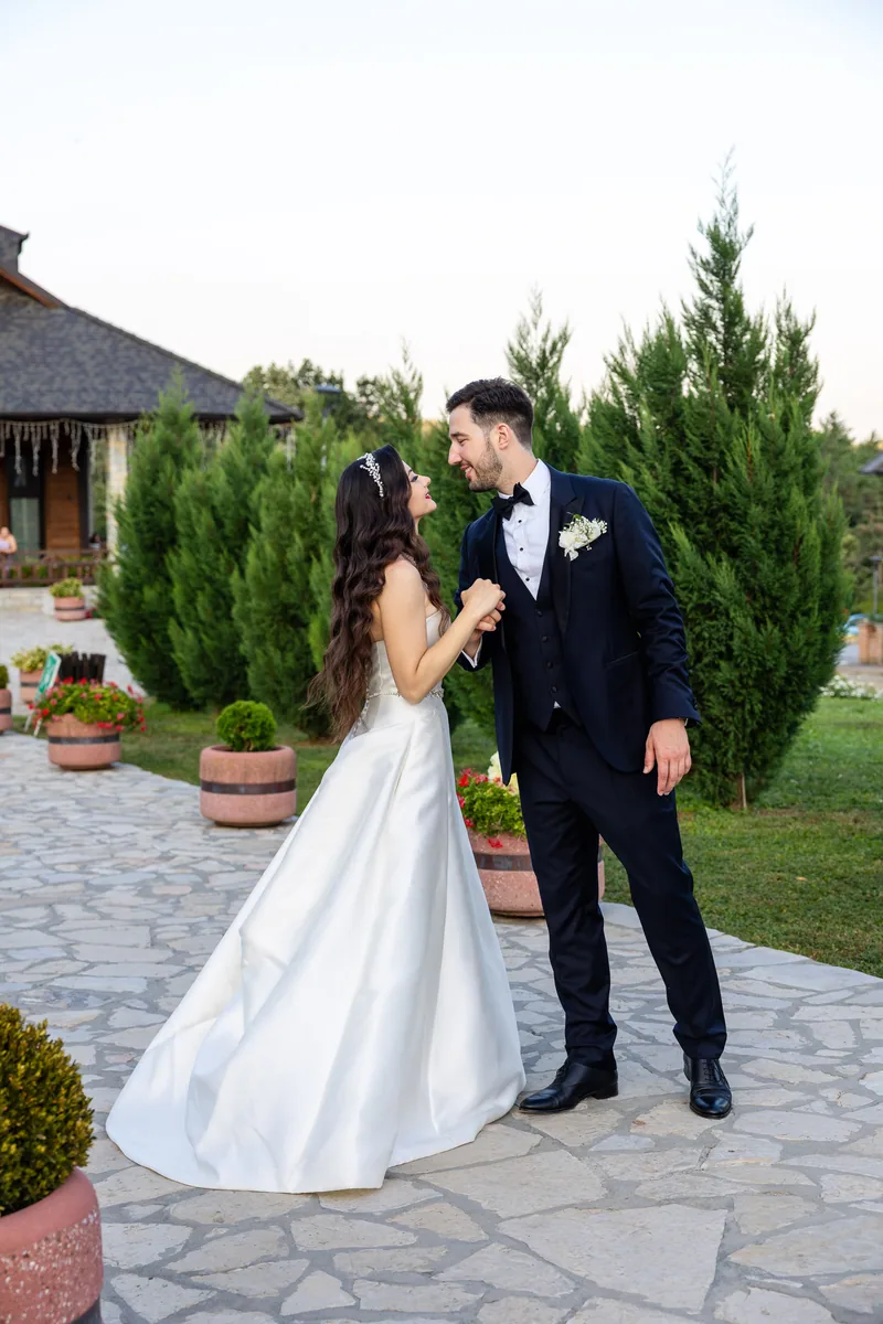 Bride and groom hold hands in a garden at dusk framed by tall cypress trees.