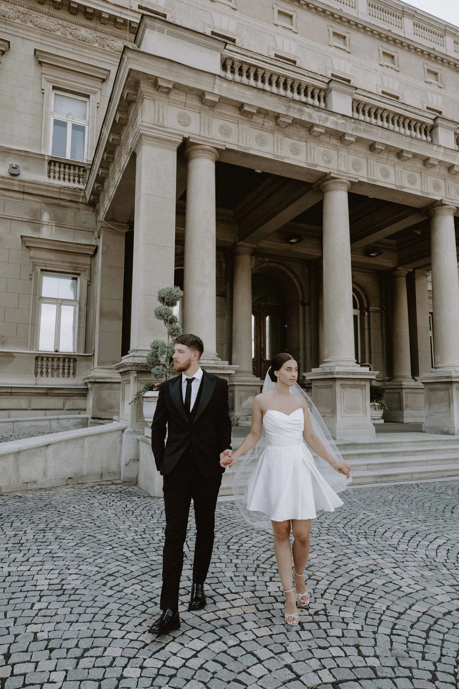 A couple hold hands walking across cobblestones before a grand columned building, the bride in a short dress.