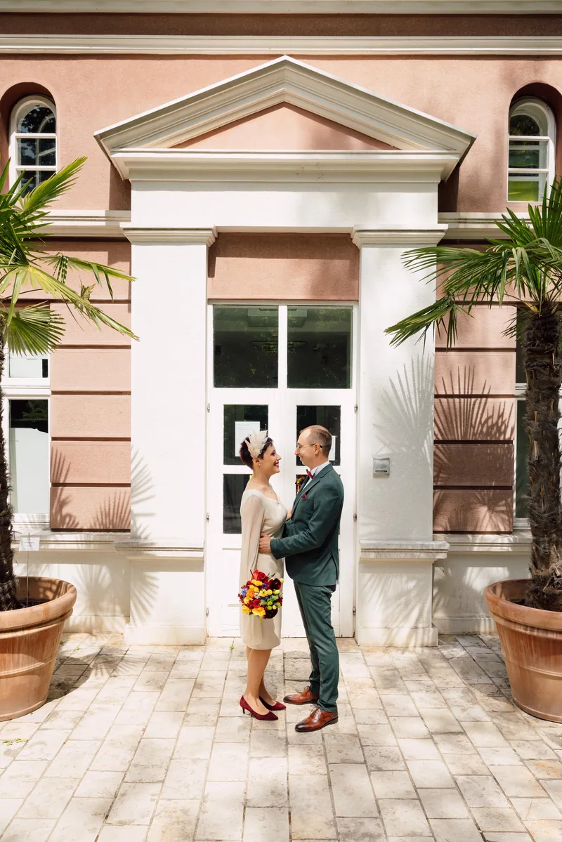 Bride and groom face each other before a pink building entrance flanked by potted palm trees