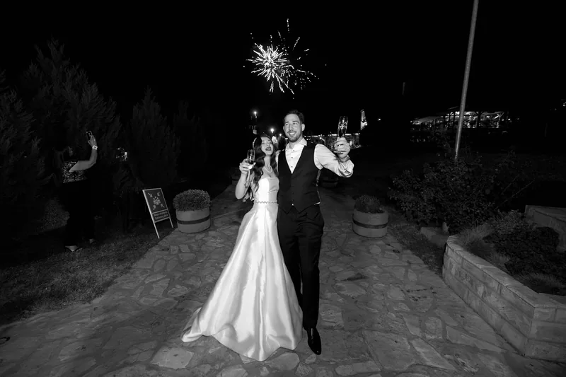 Couple walk a garden path raising glasses as fireworks burst in the night sky.