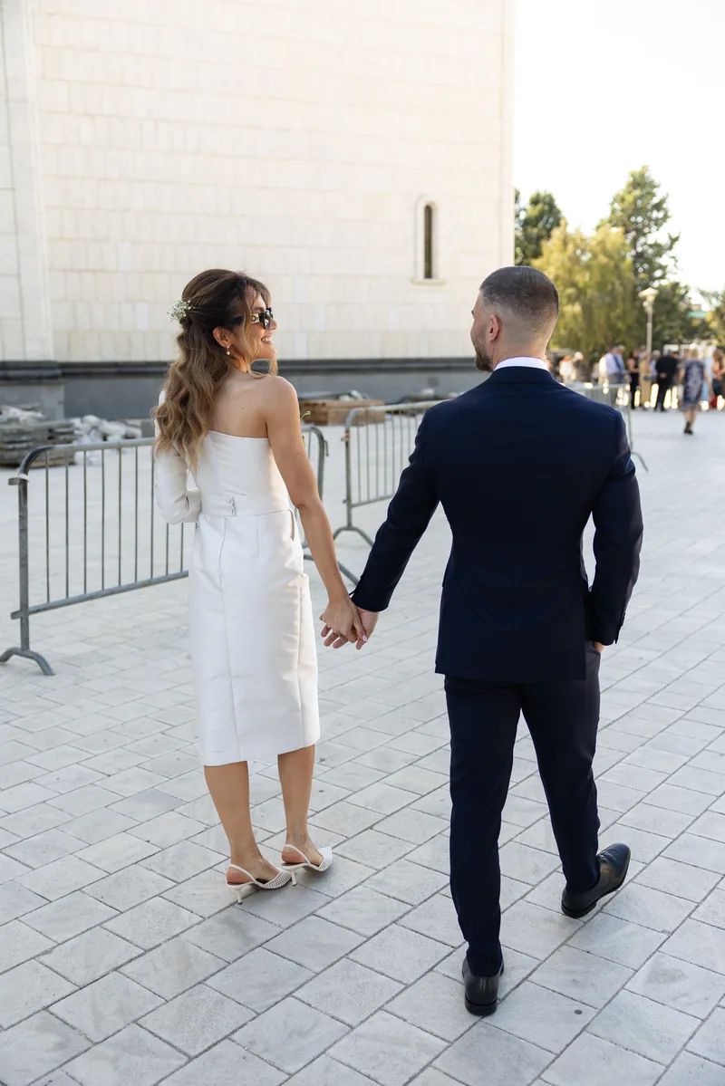 Couple stroll across a stone plaza as the bride glances back over her shoulder in sunglasses.