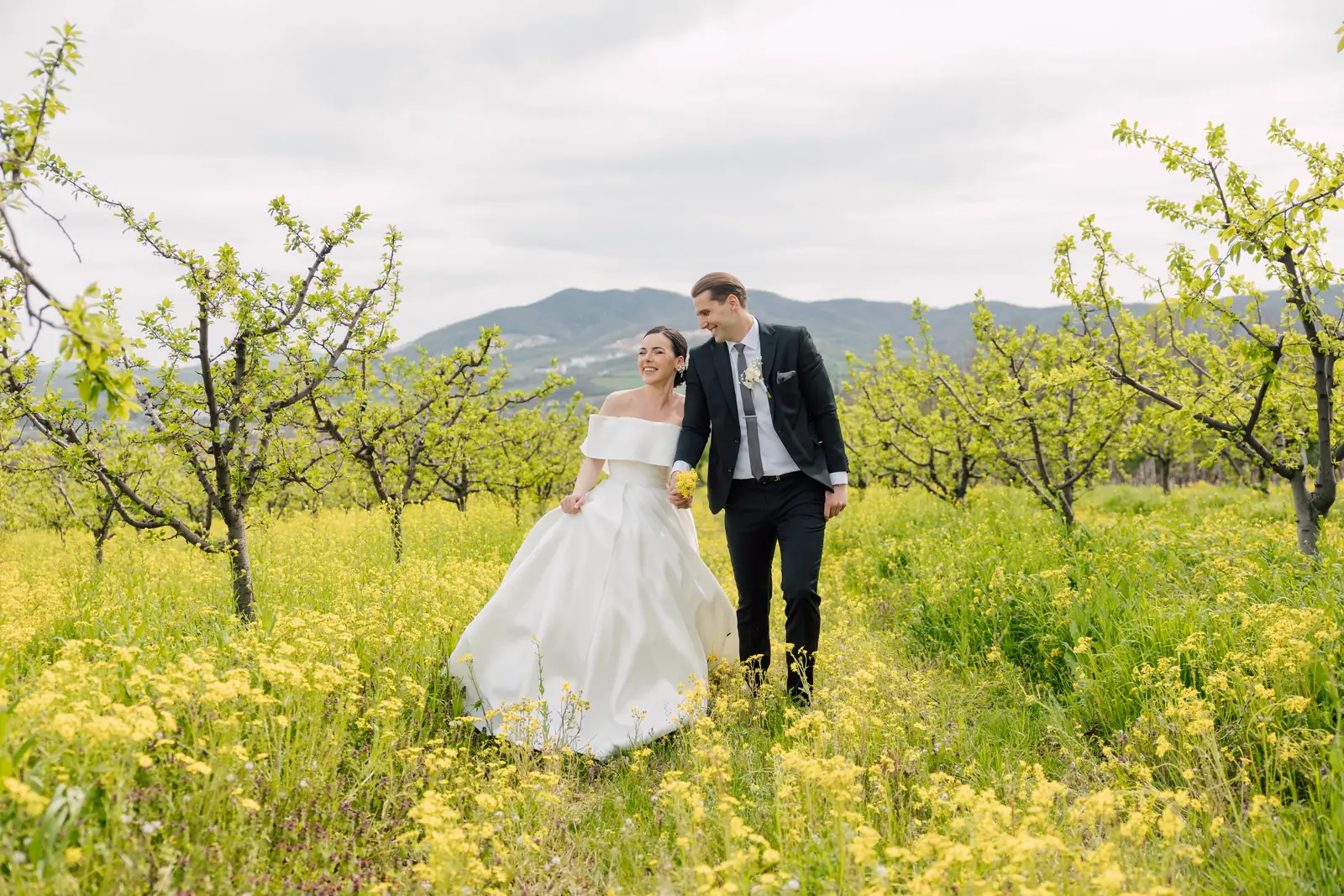 A bride and groom walk hand in hand through a yellow wildflower meadow with wooded hills rising behind them.