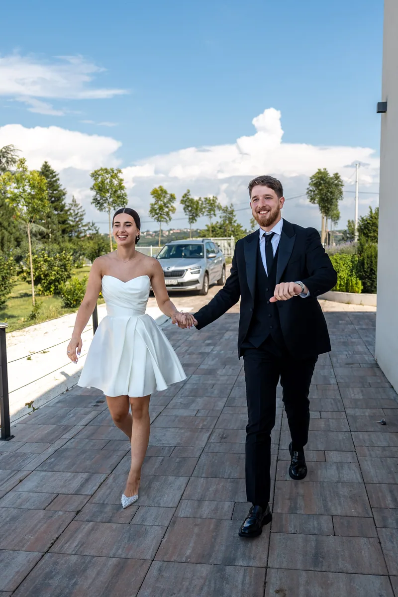 A smiling couple walk hand in hand across a wooden terrace under a bright blue summer sky.