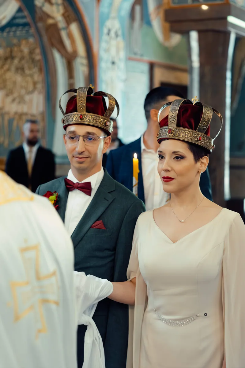 Crowned bride and groom stand side by side as a priest in gold vestments faces them