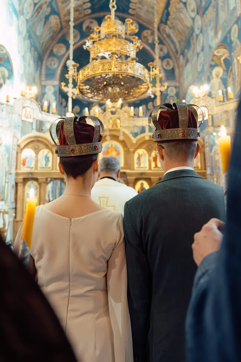 Rear view of a crowned bride and groom facing the gold altar beneath a large chandelier