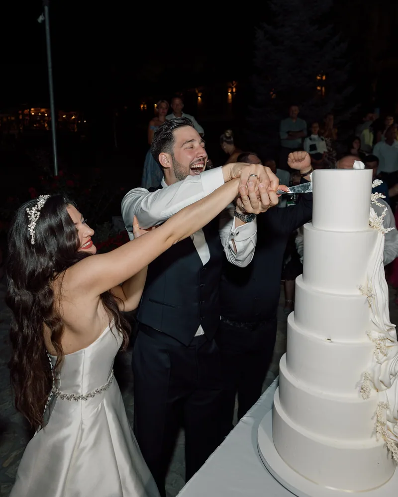 Bride and groom laugh while cutting a tall tiered white wedding cake at night.