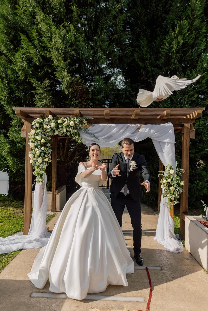 A bride and groom release a white dove that flies above a wooden pergola dressed with white flowers.