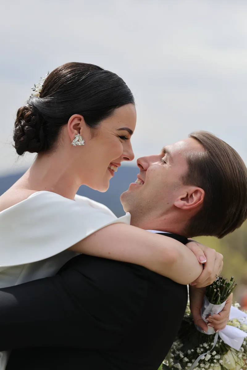 A couple smile closely face to face as the bride wraps her arms around the groom holding a white bouquet.