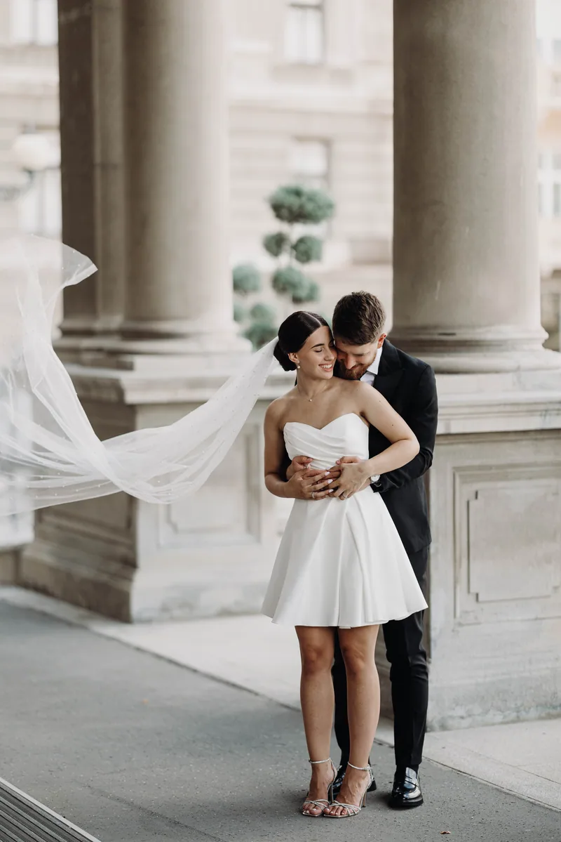 A groom holds the bride from behind on a sidewalk as her long veil streams sideways past stone columns.
