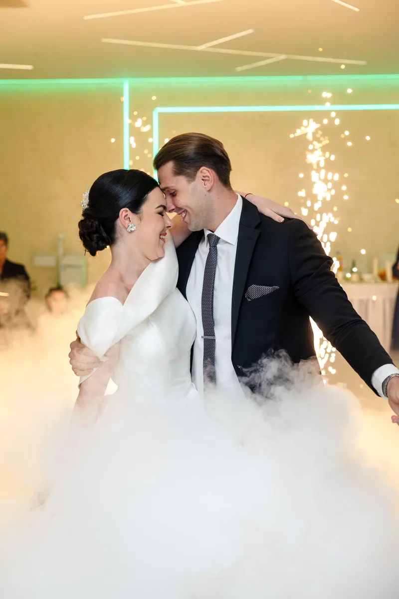 A bride and groom touch foreheads and smile during their first dance as sparklers glow behind them.