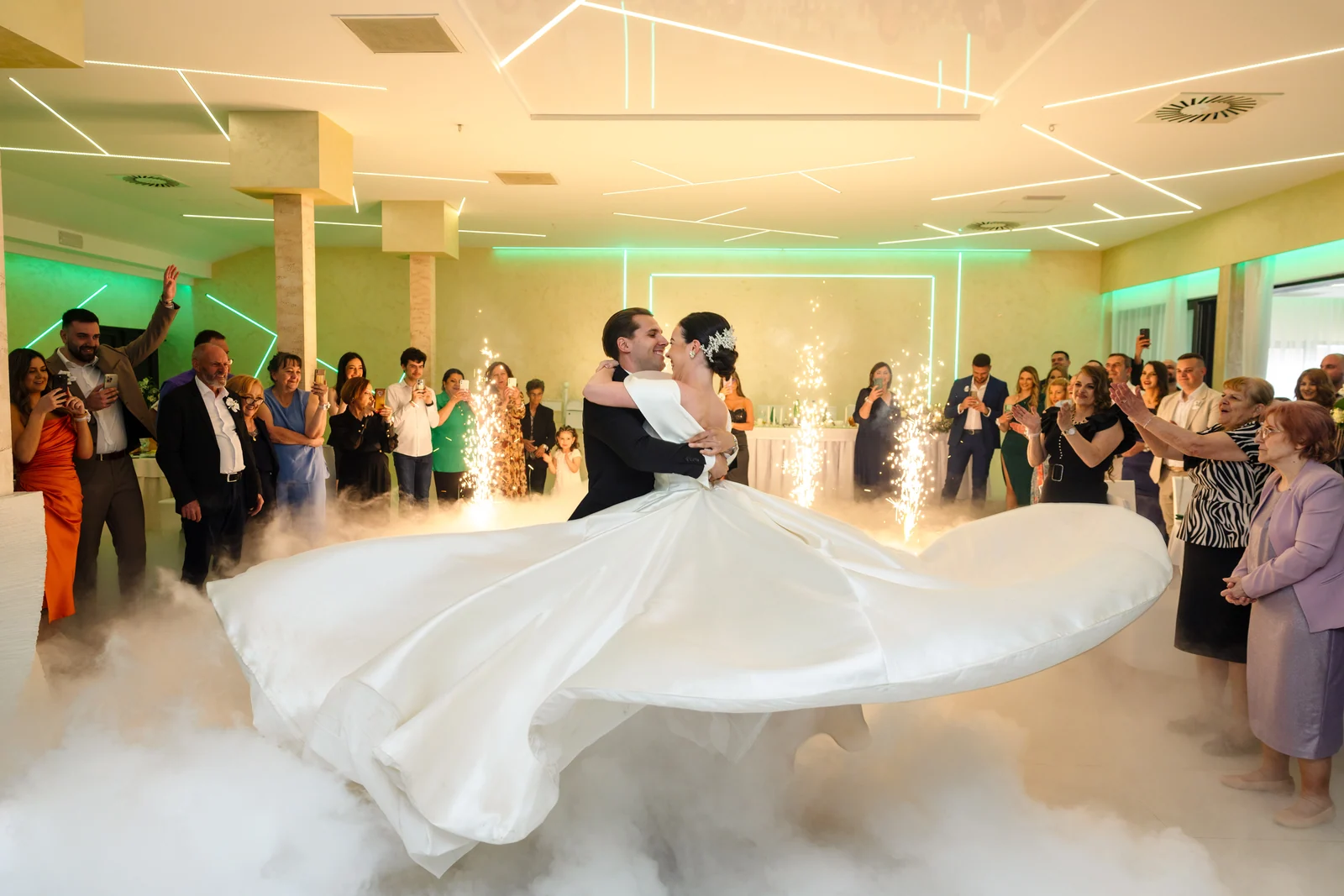 A couple share a first dance amid low fog and sparkler fountains as guests watch in a green-lit hall.