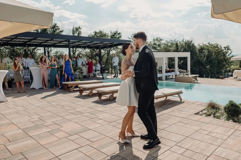 A couple share their first dance on a poolside terrace as guests watch from the shade behind them.