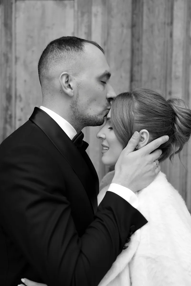 In black and white, a groom kisses his smiling bride on the forehead while cradling her face.