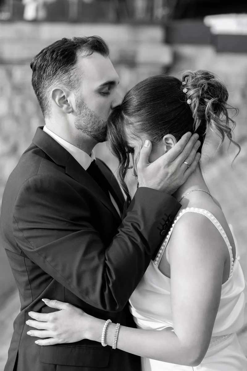 In black and white, a groom kisses his bride's forehead while gently cradling her head near stone steps.