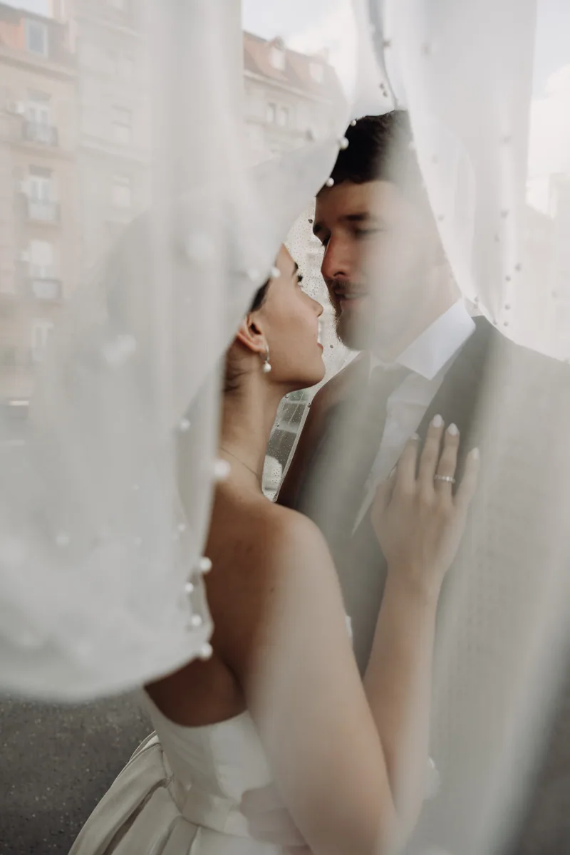 A bride and groom press their foreheads together framed by a flowing pearl veil, city buildings behind them.