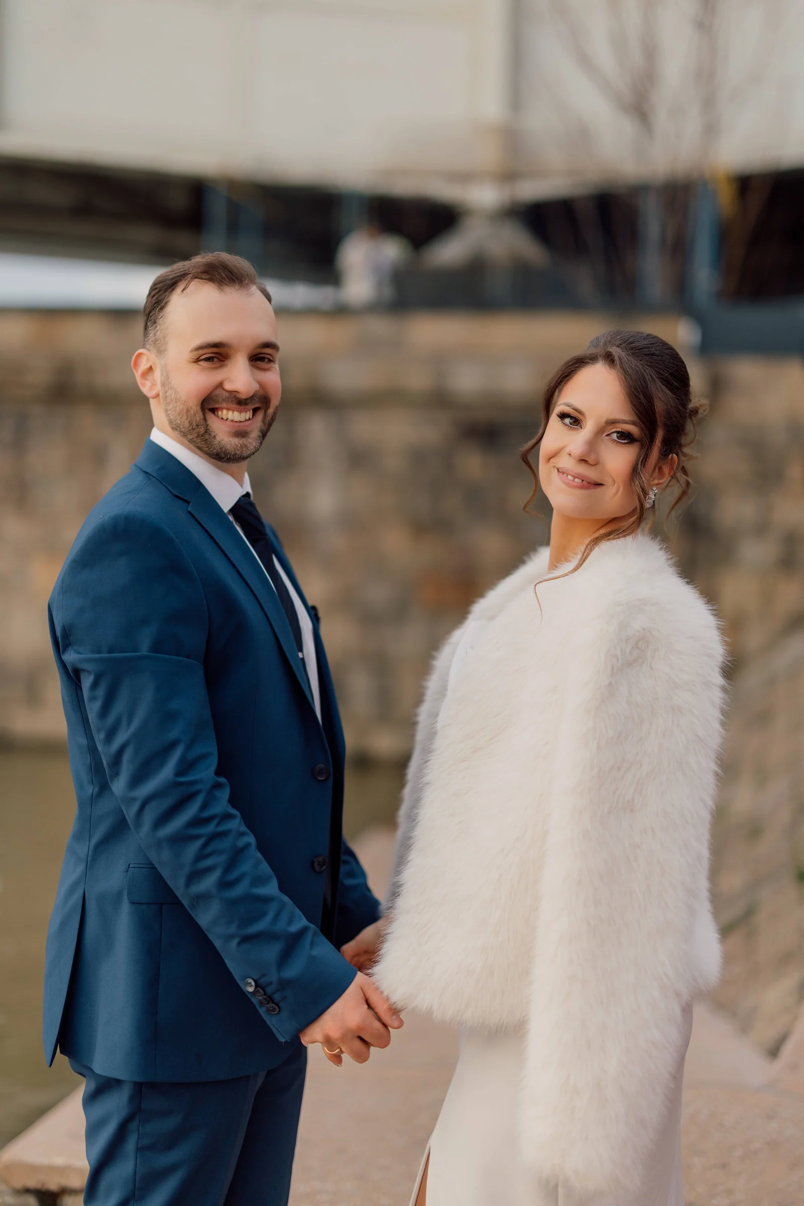 A smiling couple hold hands by a stone river embankment, the bride wearing a soft white fur coat.