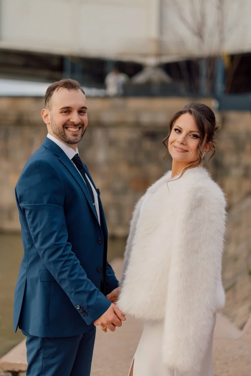A smiling couple hold hands by a stone river embankment, the bride wearing a soft white fur coat.
