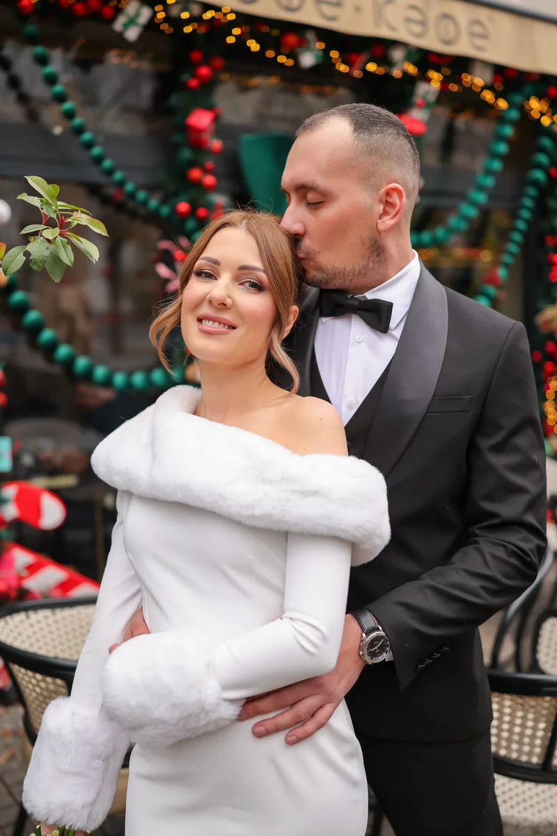 A groom kisses the head of his bride, who wears a white fur stole, in front of a cafe decorated for Christmas.