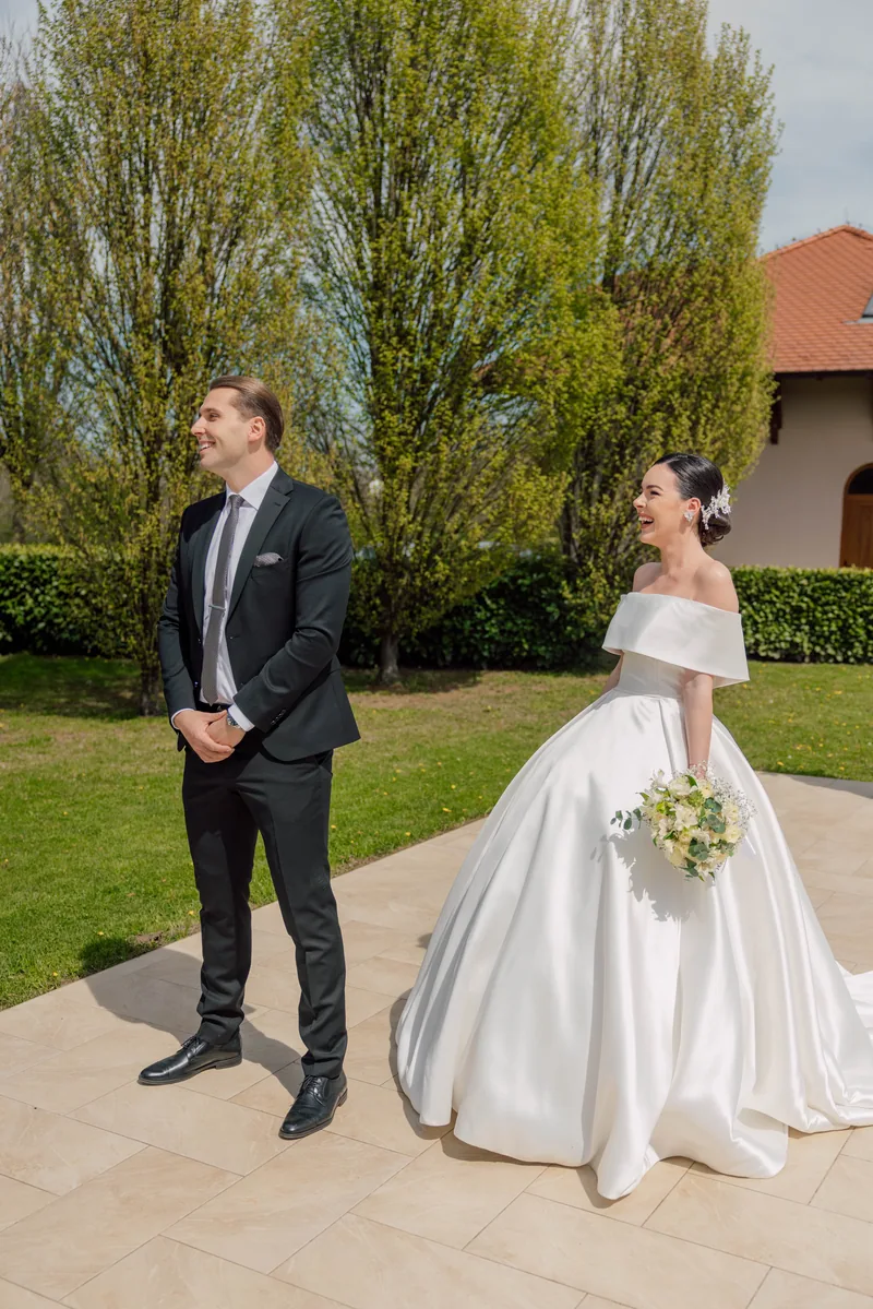 A groom waits as his laughing bride approaches with a bouquet across a garden path lined with spring trees.