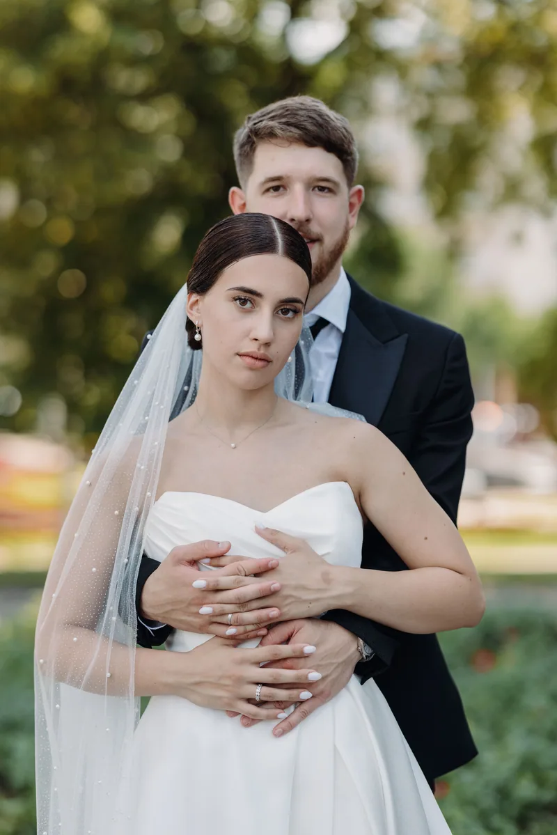 A groom wraps his arms around the bride from behind in a green garden, both looking calmly at the camera.