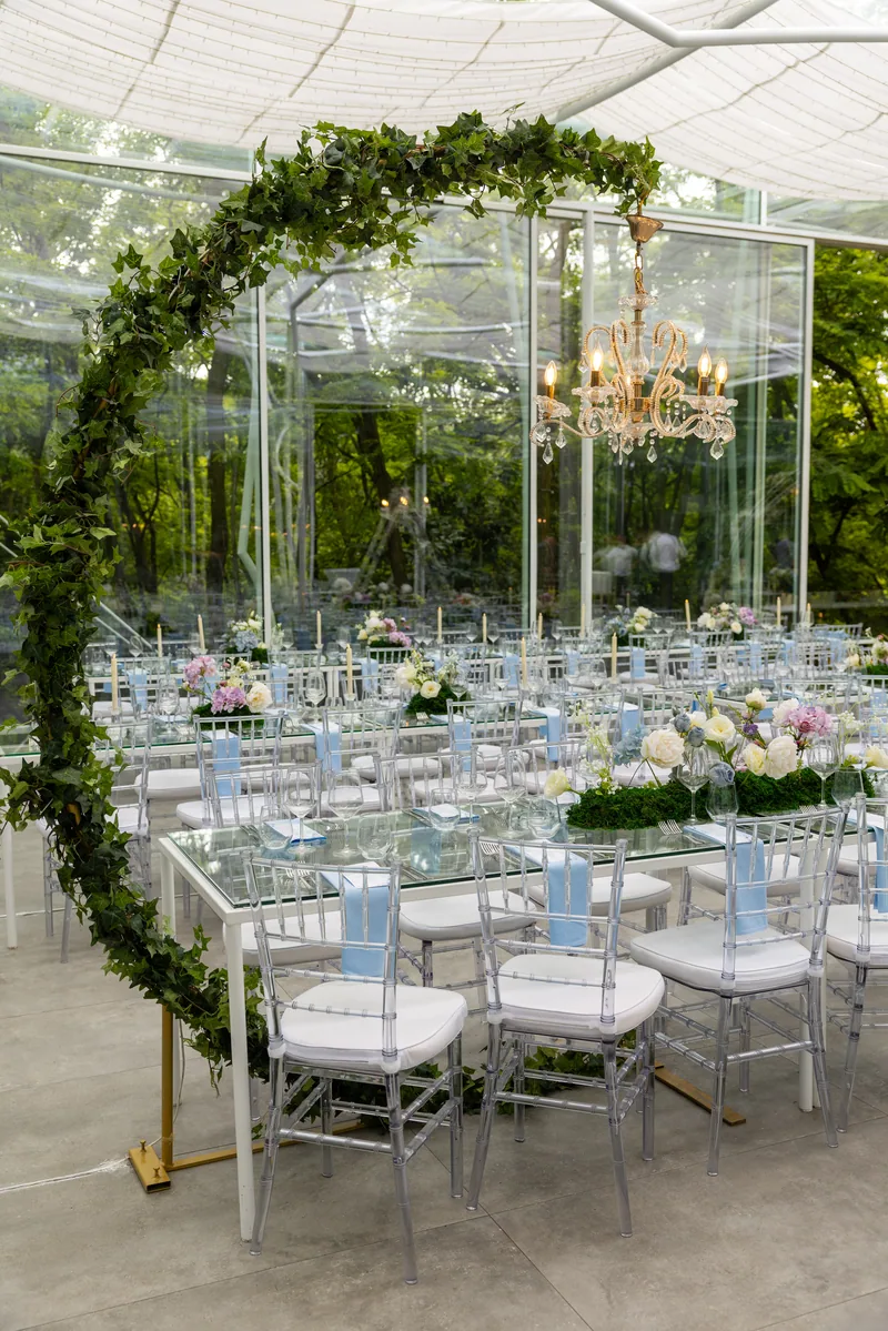 Glass pavilion reception set with an ivy arch, crystal chandelier, clear chairs and pastel flowers