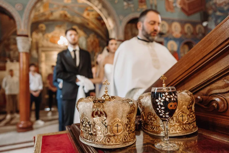 Two golden ceremonial crowns and a glass of wine rest on a table as a priest and couple stand behind.