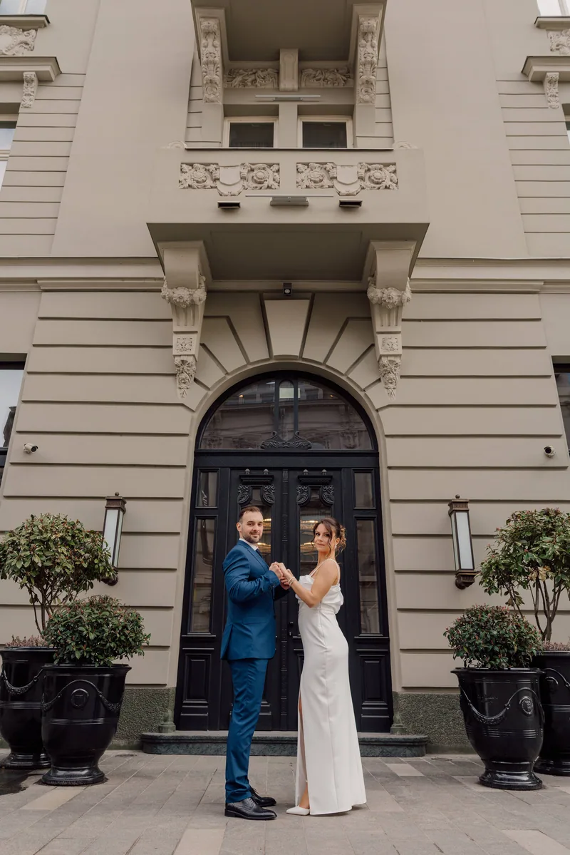 A couple hold hands and face the camera before a grand arched doorway flanked by two potted shrubs.